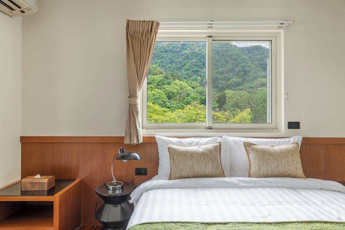 Une chambre d’hôtel dotée d’une grande fenêtre, d’une tête de lit en bois et d’une table de chevet avec une lampe.