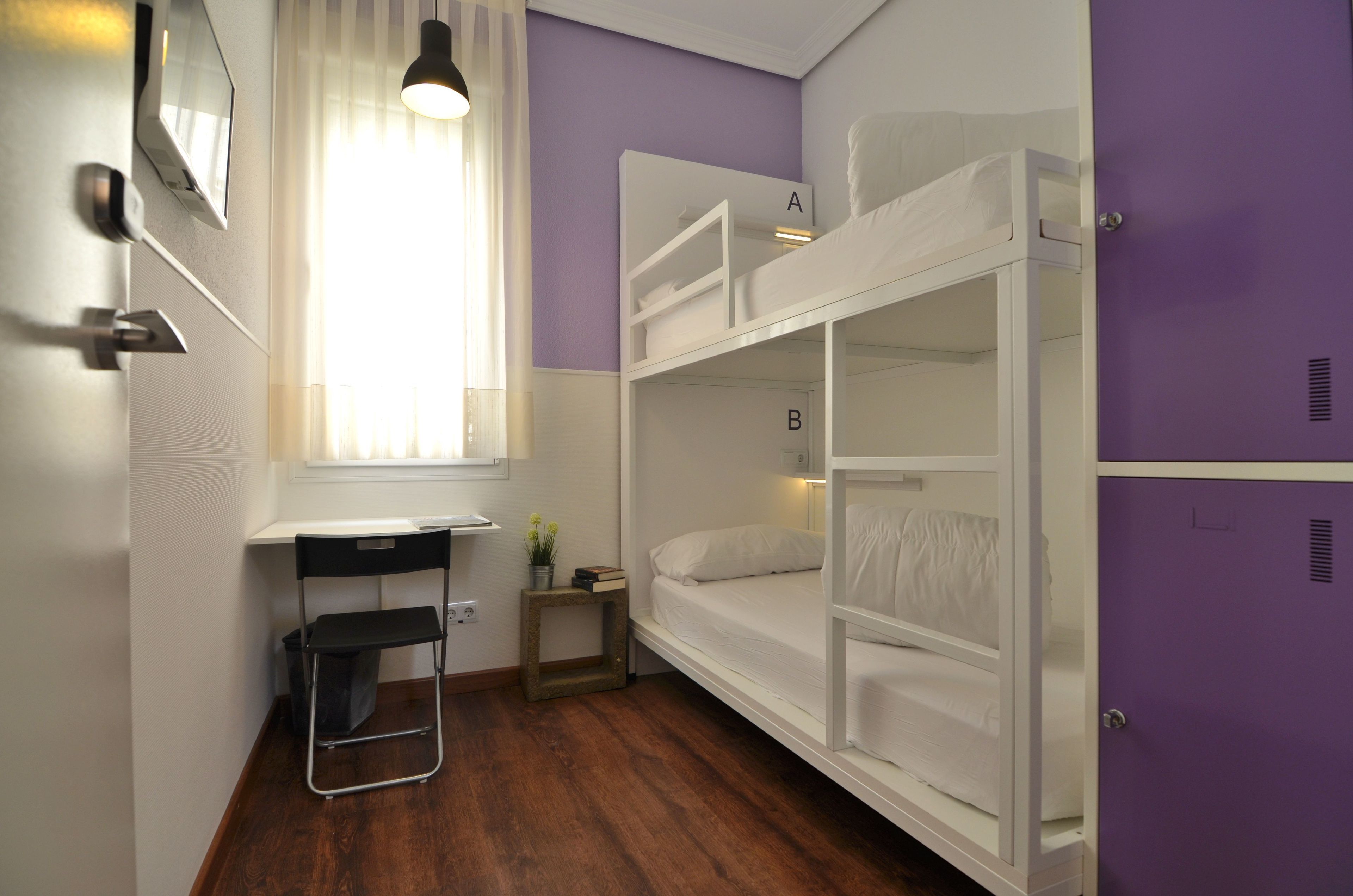 A dormitory room with bunk beds labeled A and B, a desk with a chair, a small table with a plant, and a window with curtains.