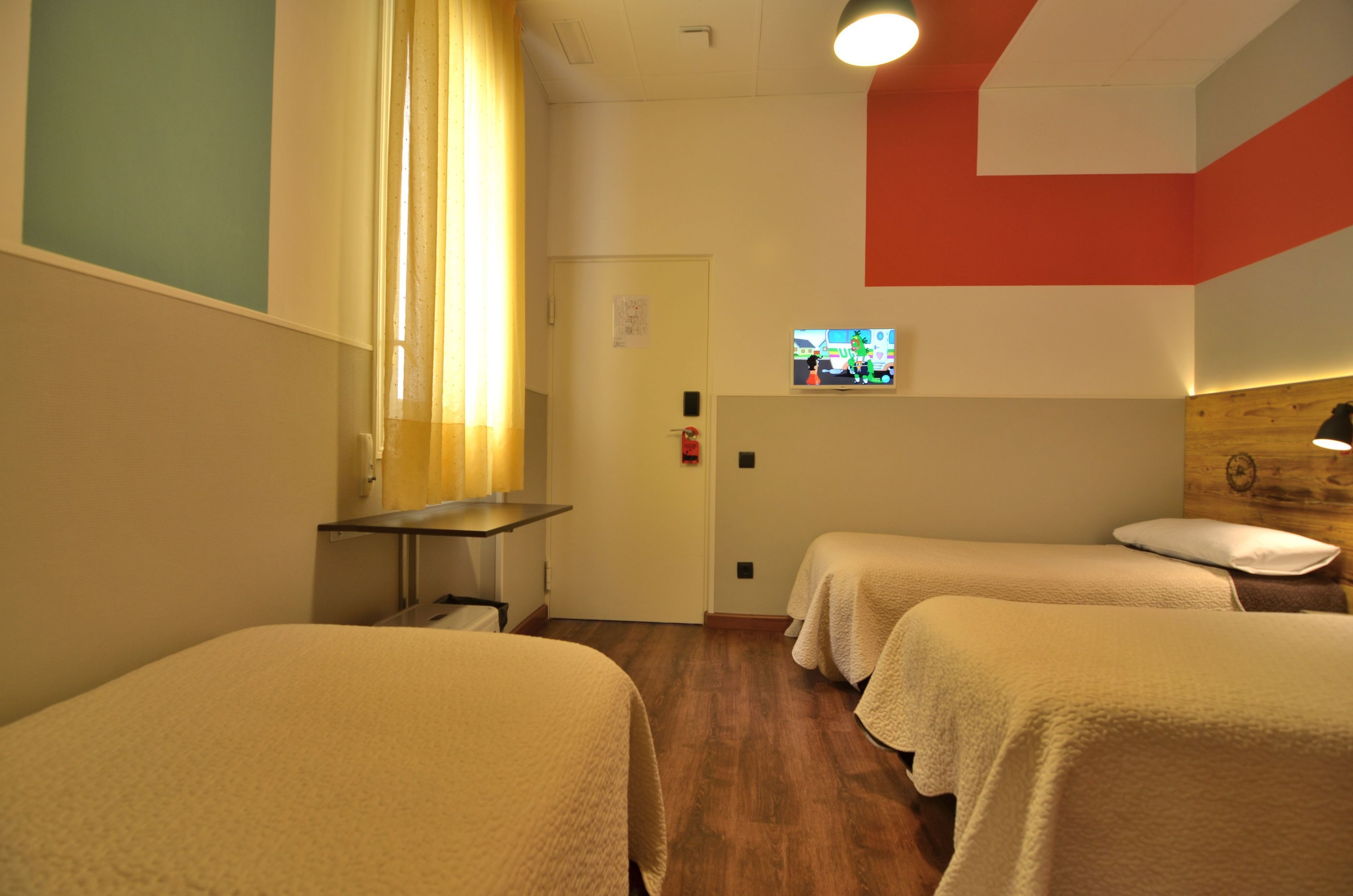 A room with two beds, a small table, a TV on the wall, and a wooden accent wall.