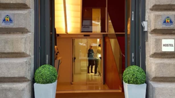 A hostel entrance with a glass door, two potted plants, and a sign indicating the hostel.