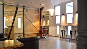A spacious room with wooden flooring, a large window, and a kitchen area visible through an open door.