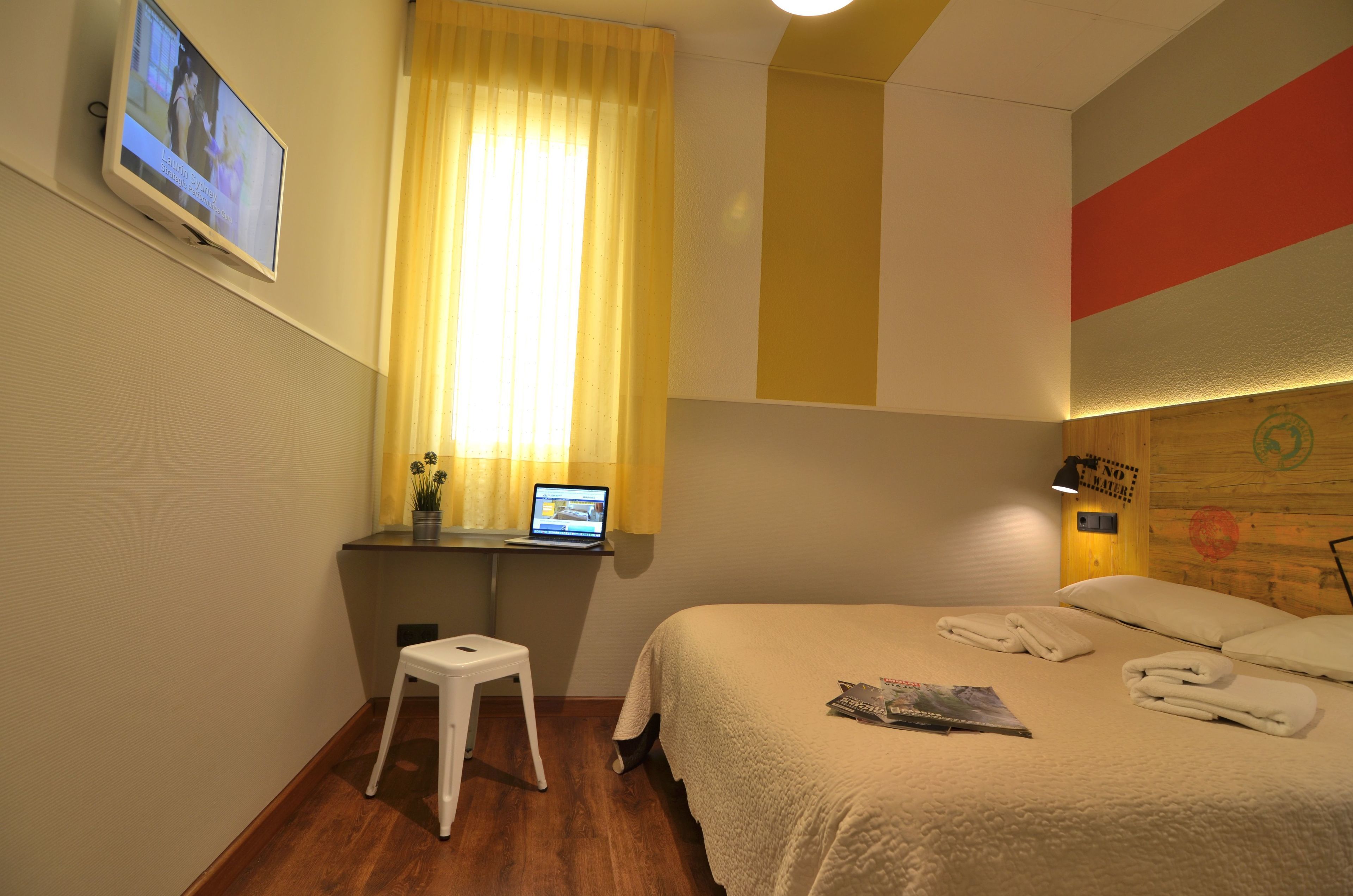 A hotel room with a bed, a desk with a laptop, a TV mounted on the wall, and a window with curtains.