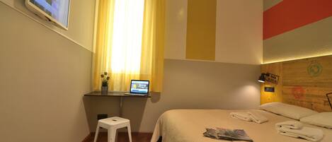 A hotel room with a bed, a desk with a laptop, a TV mounted on the wall, and a window with curtains.