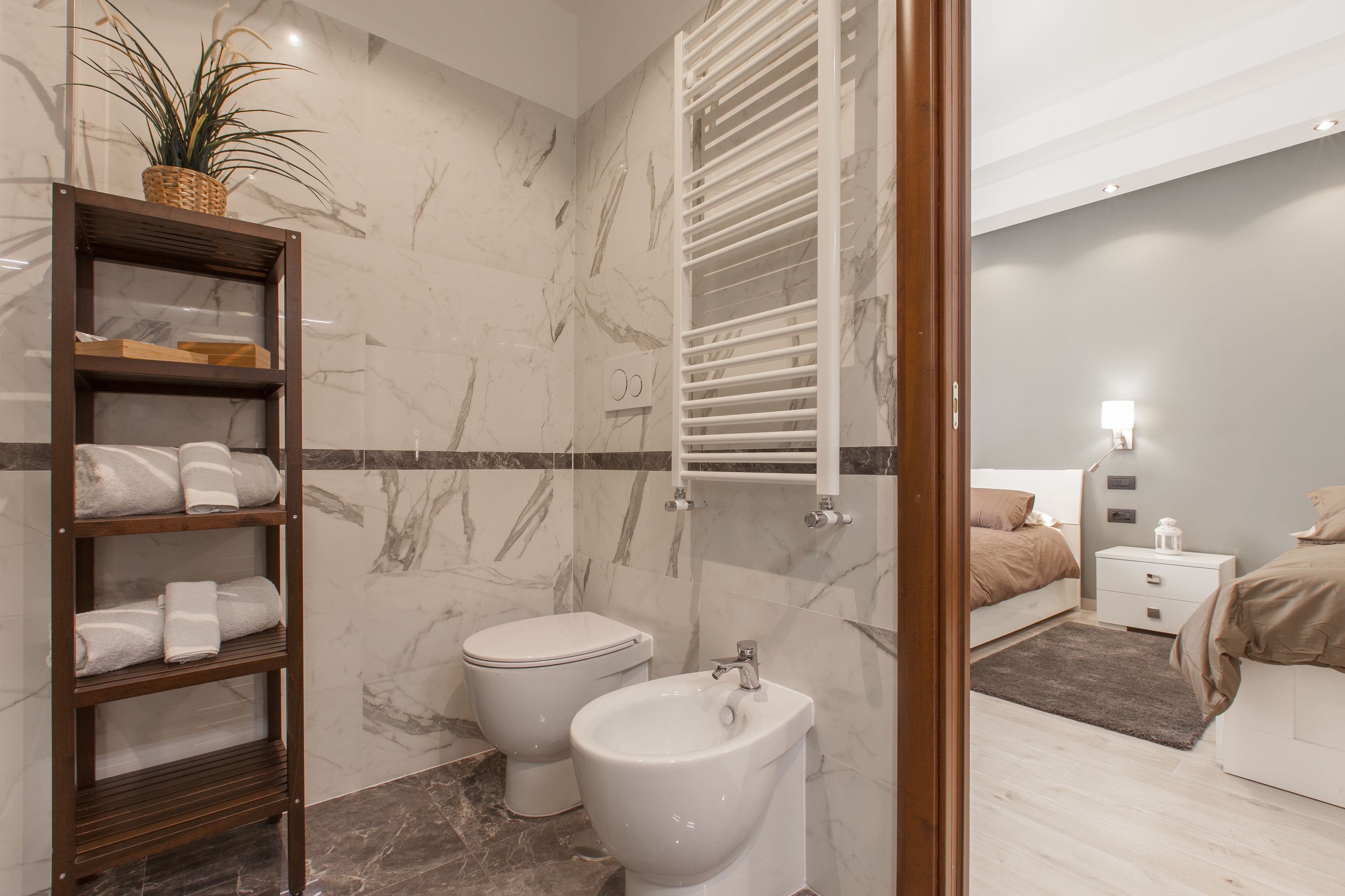 Une salle de bain moderne avec un mur en marbre, une étagère en bois où sont posées des serviettes, des toilettes, un bidet et une table de chevet avec une lampe.