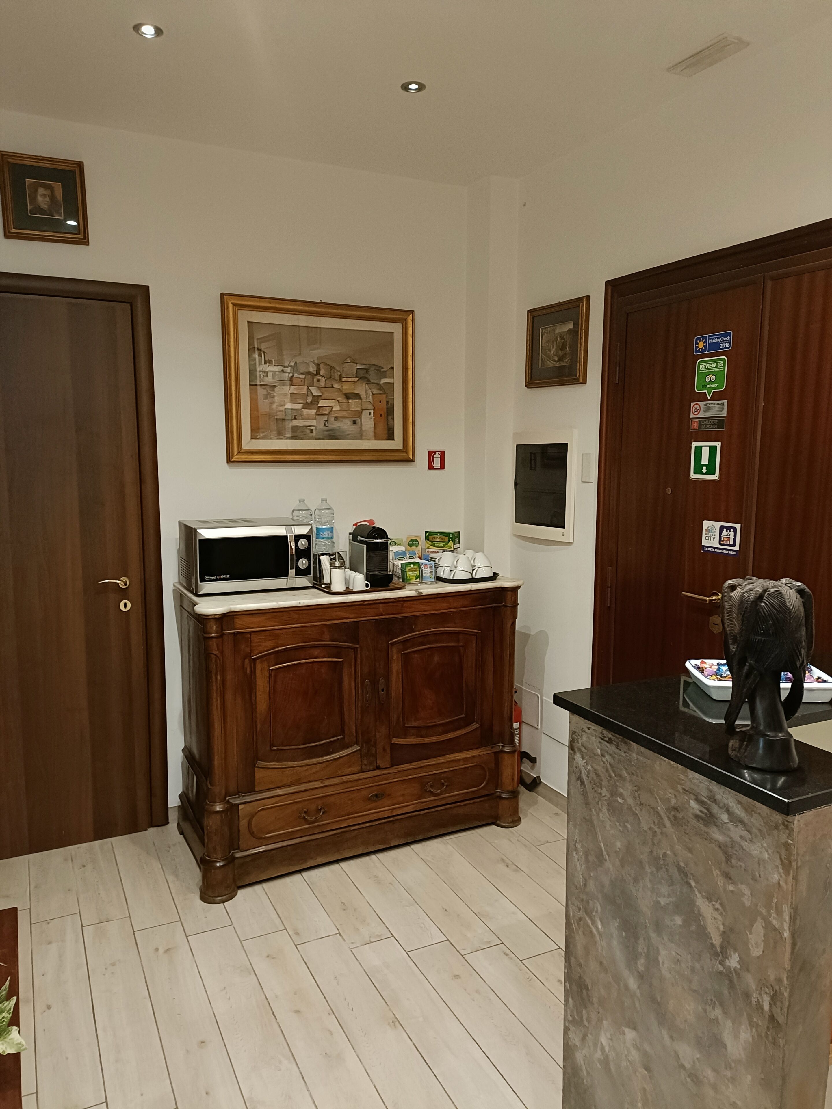 Een keuken met een houten kast, een magnetron en een koffiezetapparaat. Er hangen ingelijste foto's aan de muur en is een deur met een bordje.