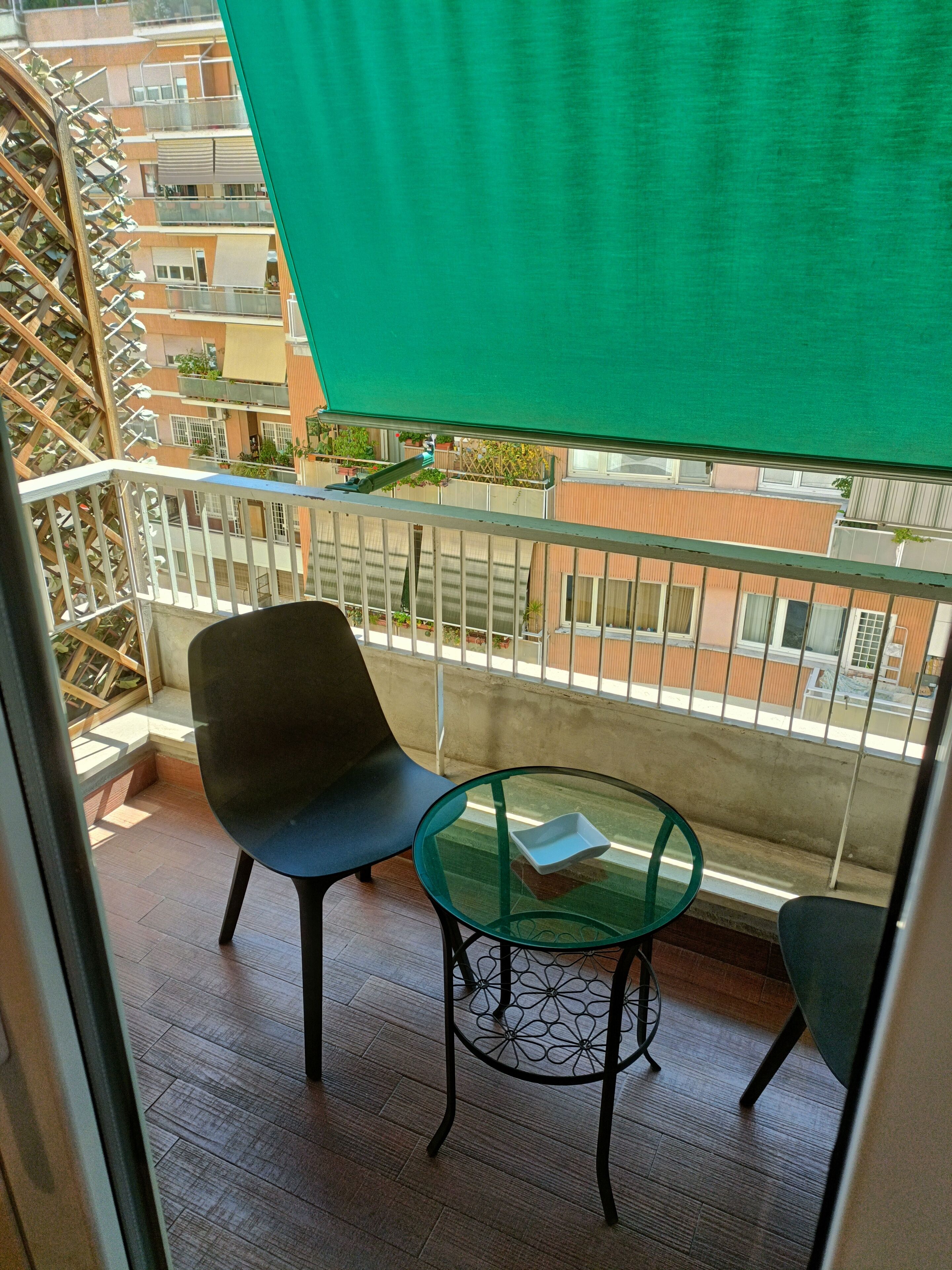 A balcony with a glass table and two chairs, a green sunshade, and a view of neighboring buildings.