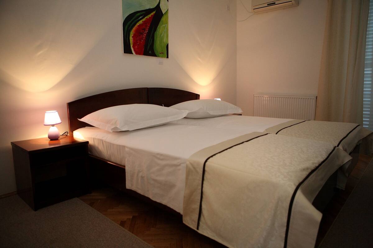 A neatly made bed with white linens and a dark headboard, a bedside lamp, and a wall-mounted air conditioner.