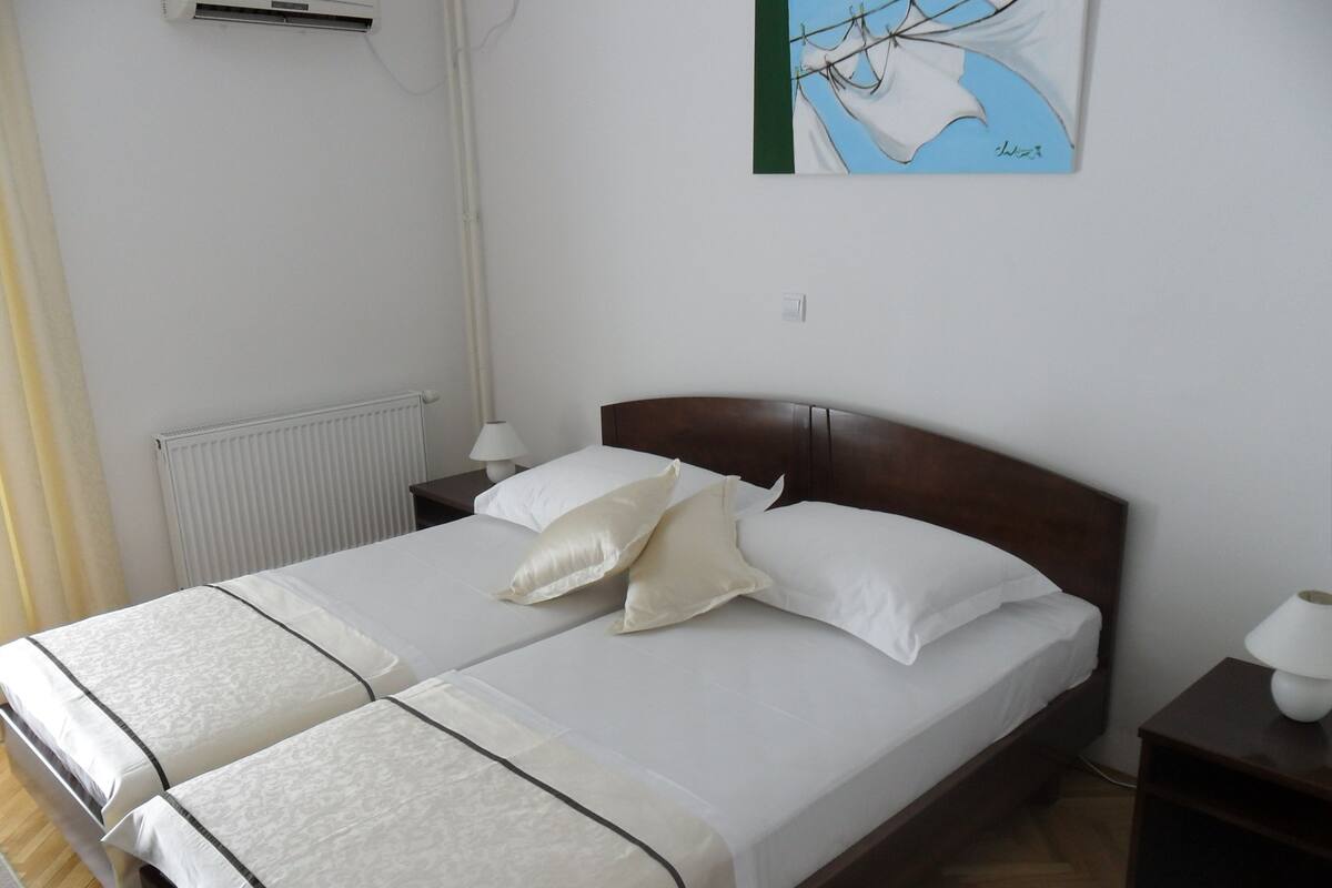 A double bed with white linens and two pillows, a bedside table with a lamp, a radiator, and a painting on the wall.