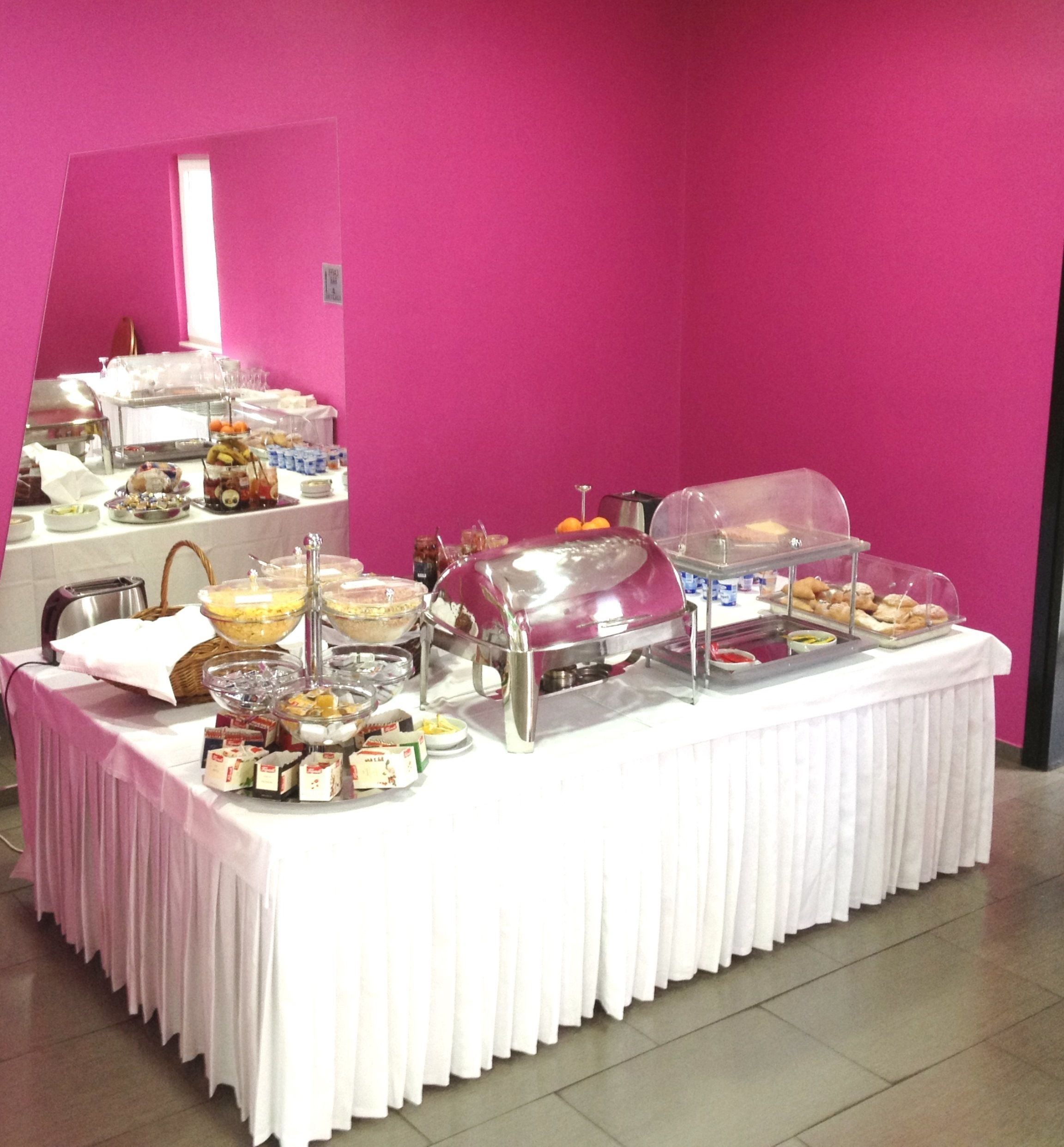Een buffettafel met diverse etenswaren en een zelfbedieningsdrankstation, voor een roze muur.