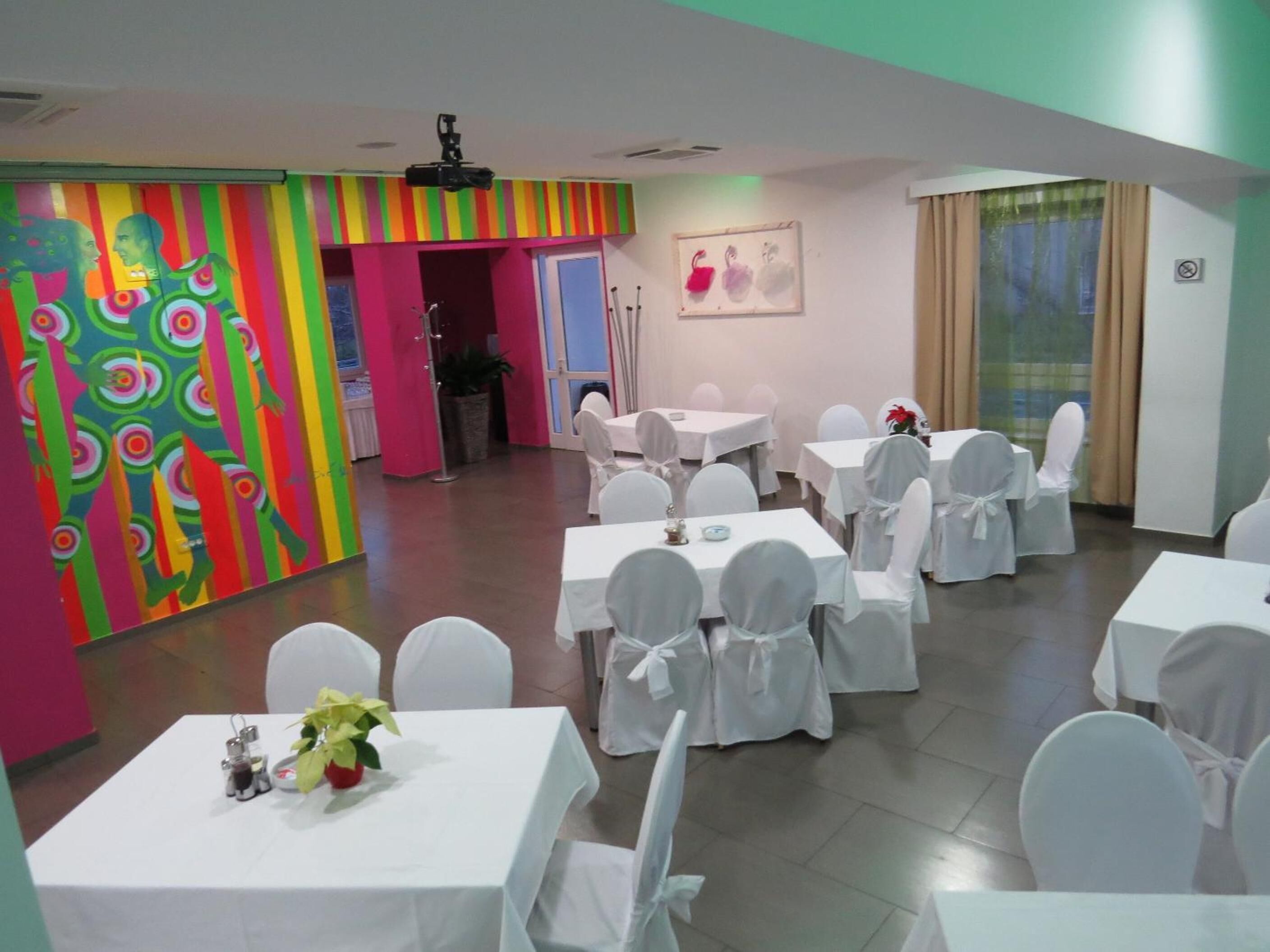 A conference room with white tables and chairs, a colorful mural, and a projector on the ceiling.