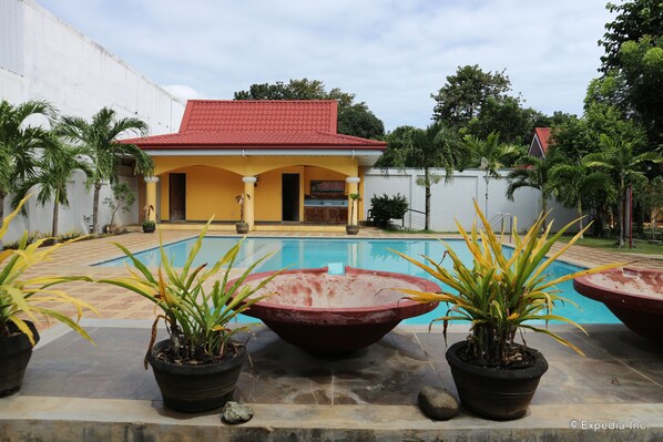A swimming pool with a red roof building in the background, surrounded by potted plants.
