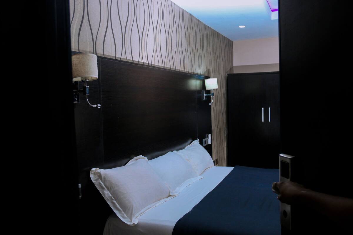 Une chambre d’hôtel avec un grand lit, une tête de lit en bois foncé, deux lampes fixées au mur et une armoire.