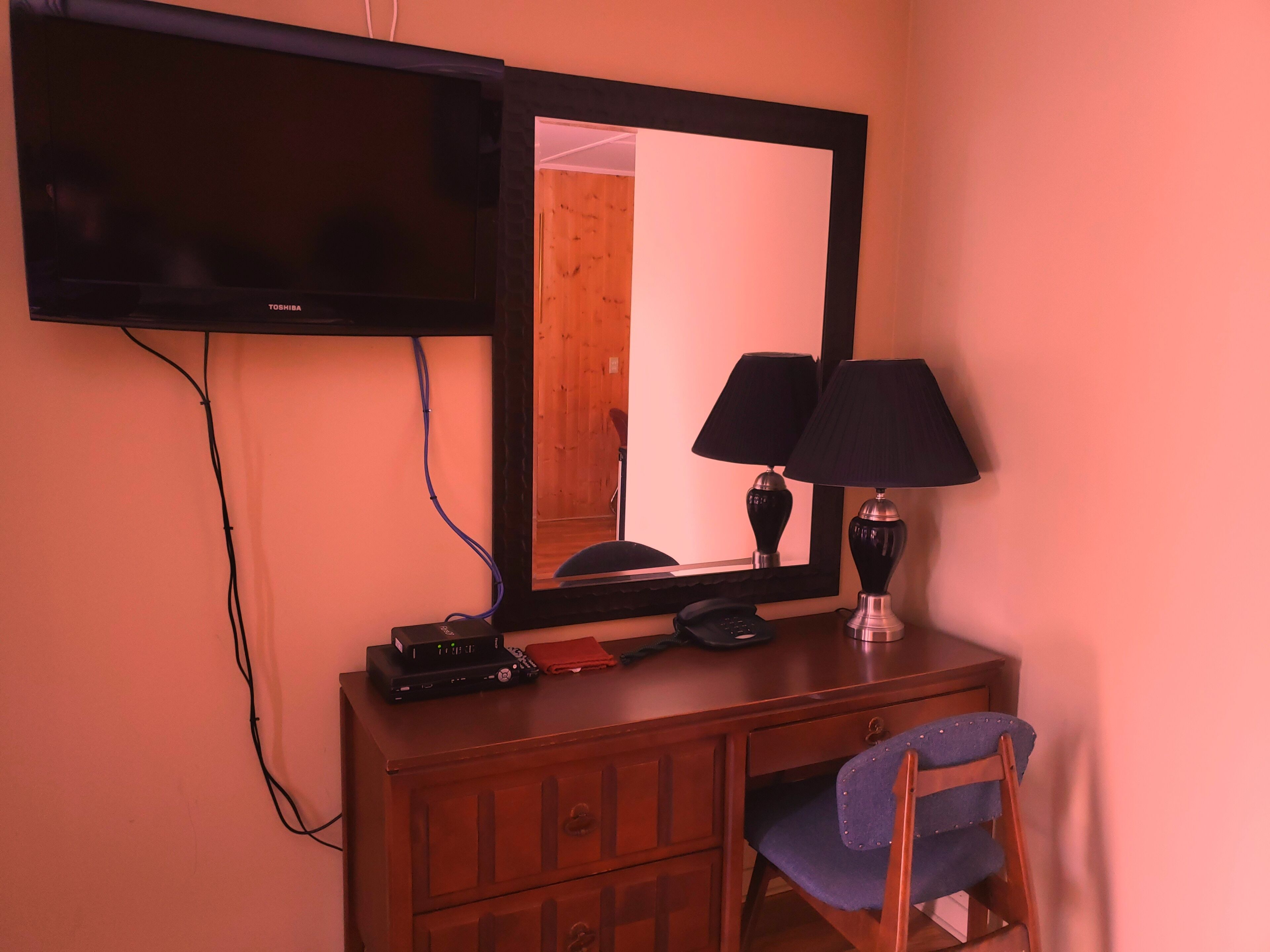 A room with a television mounted on the wall, a desk with a telephone, a lamp, and a chair.