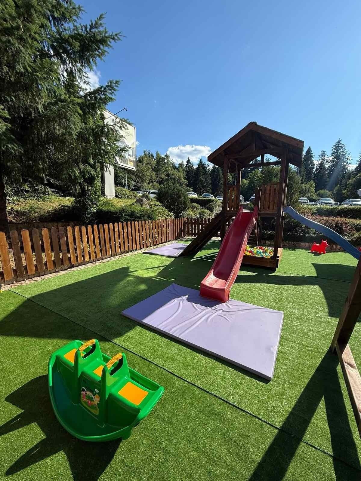 A playground with a slide, a small wooden structure, and artificial grass.