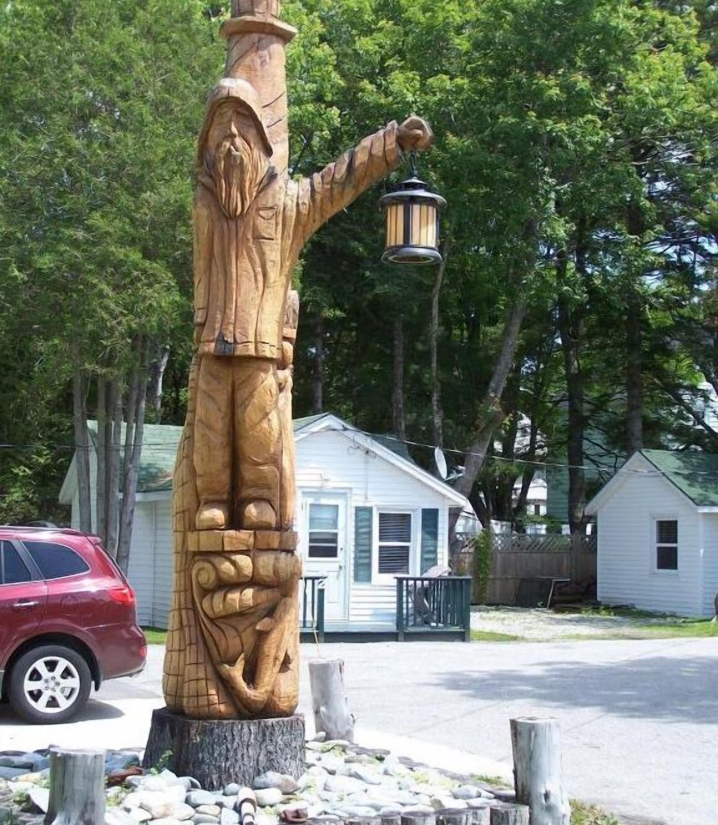 Une sculpture en bois représentant une figure tenant une lanterne, placée sur un socle en pierre et entourée d’une clôture en métal.