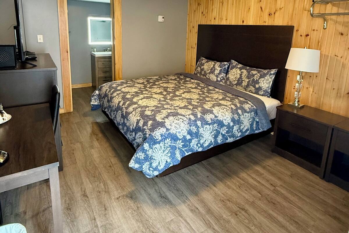 A bedroom with a wooden ceiling, a bed with a floral bedspread, a desk with a computer, and a bedside table with a lamp.