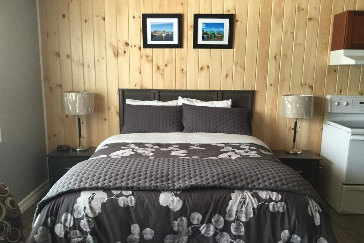 A bedroom with a wooden paneled wall, two framed pictures, a bed with a floral duvet, two bedside lamps, and a white appliance.