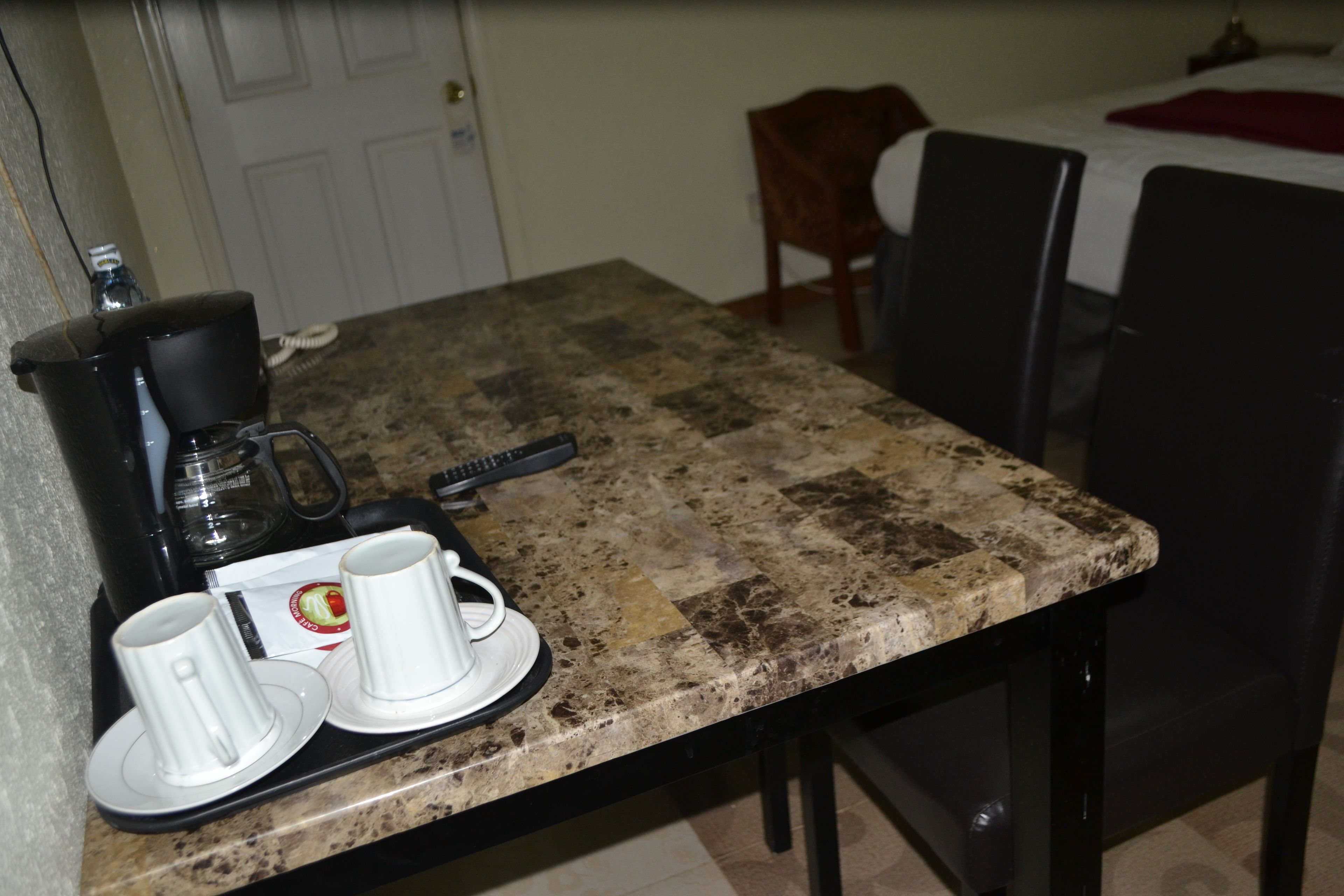 Angolo cottura con macchina del caffè, due tazze bianche e un piatto. Si vede un tavolo da pranzo con sedie e un letto sullo sfondo.