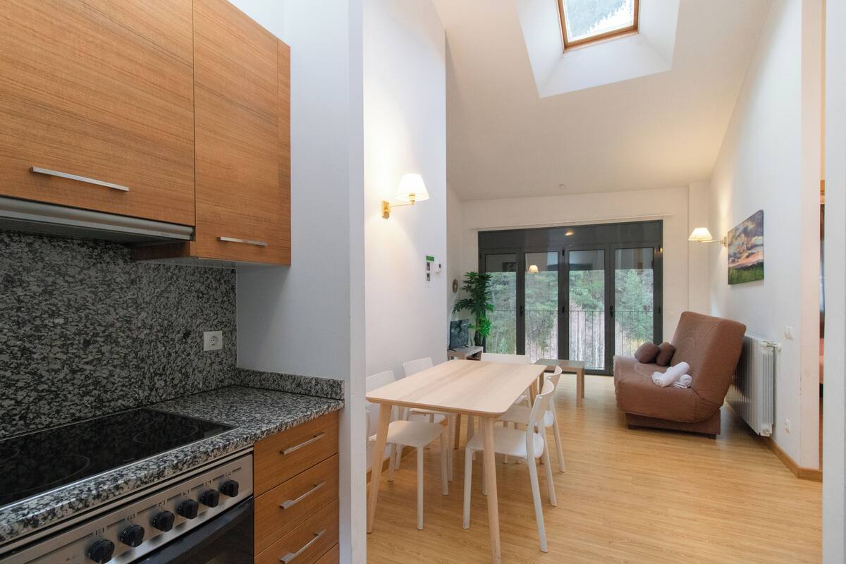 Une cuisine moderne avec des comptoirs en granit, une cuisinière encastrée et des armoires en bois. On y trouve un coin repas avec une table et des chaises, ainsi qu’un fauteuil marron avec une serviette enroulée.