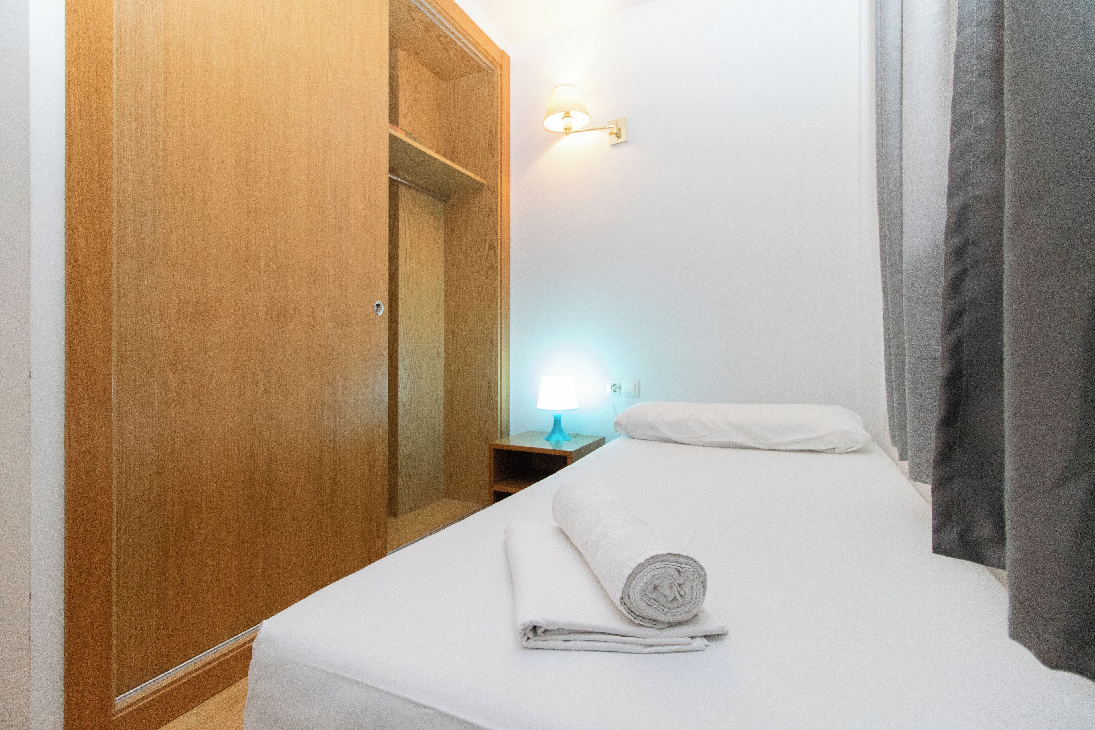 A hotel room with a wooden wardrobe, a bedside table with a lamp, a neatly made bed, and a window with curtains.