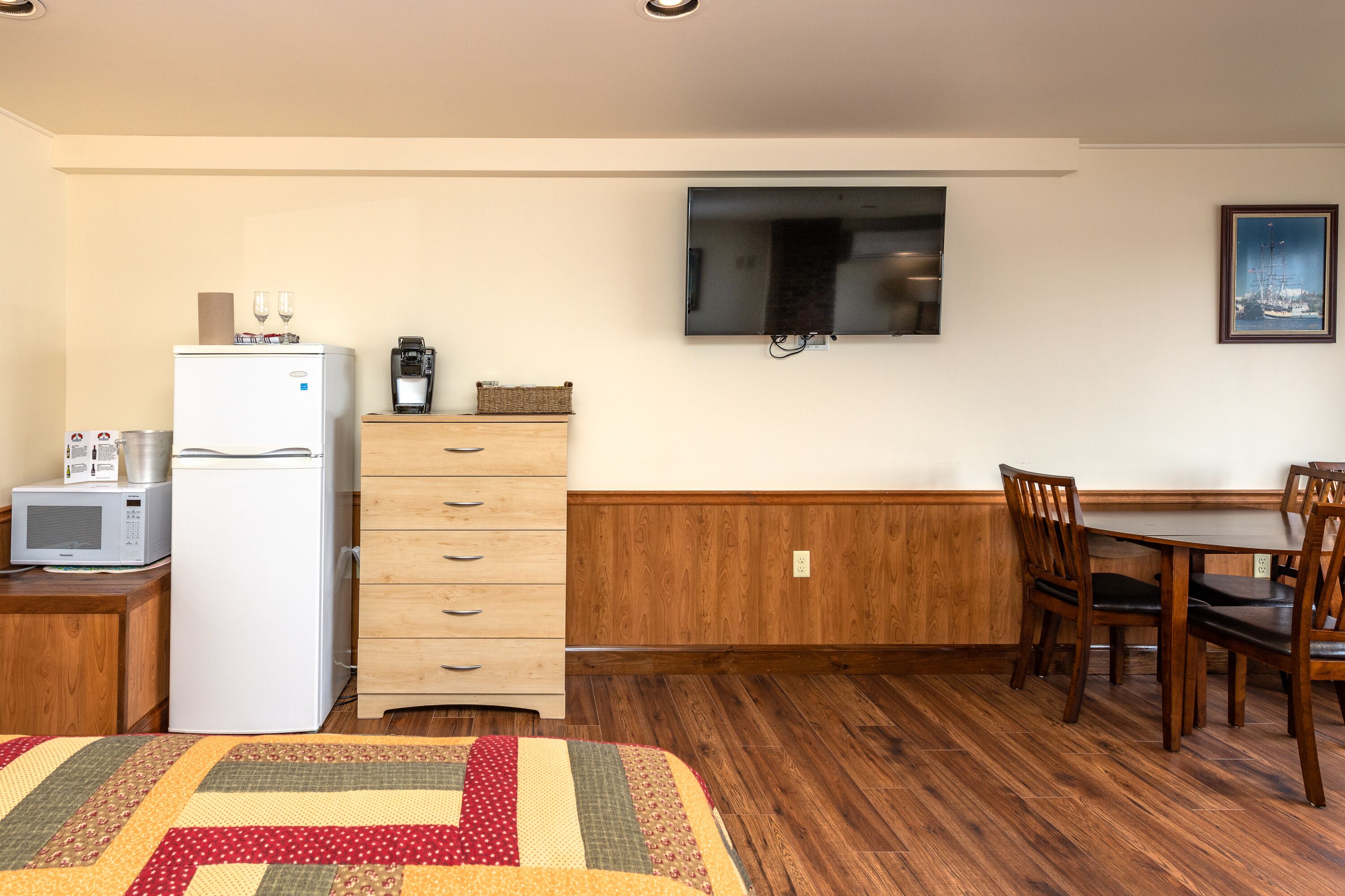 A compact room with a kitchenette, a dining area with wooden chairs, a TV mounted on the wall, and a framed picture.