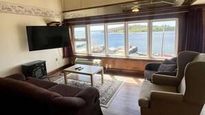 A living room with a large window overlooking a body of water, a flat-screen TV, a dark brown sofa, a patterned armchair, and a small coffee table. - The Dockside Inn & Restaurant (Lunenburg)