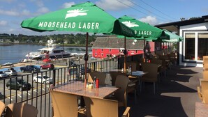 An outdoor seating area with green umbrellas labeled "Moosehead Lager," overlooking a body of water with boats and a red building in the background. - The Dockside Inn & Restaurant (Lunenburg)