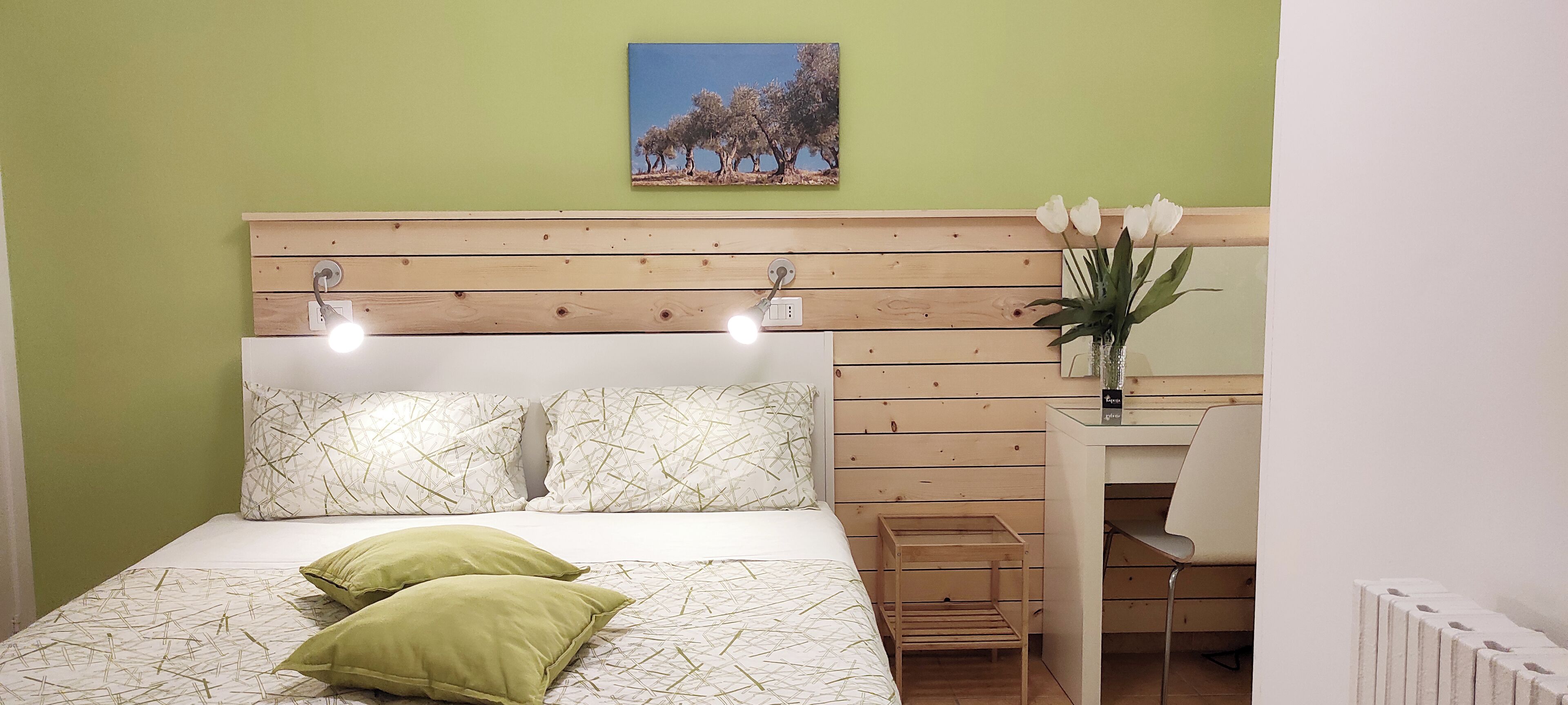 A bedroom with a wooden headboard, two bedside lamps, a bed with patterned bedding, a small bedside table with a vase of flowers, and a painting on the wall.