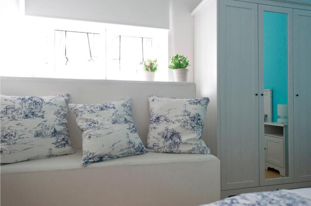 A room with a bed, a window with curtains, a wardrobe, and a bench with patterned cushions.
