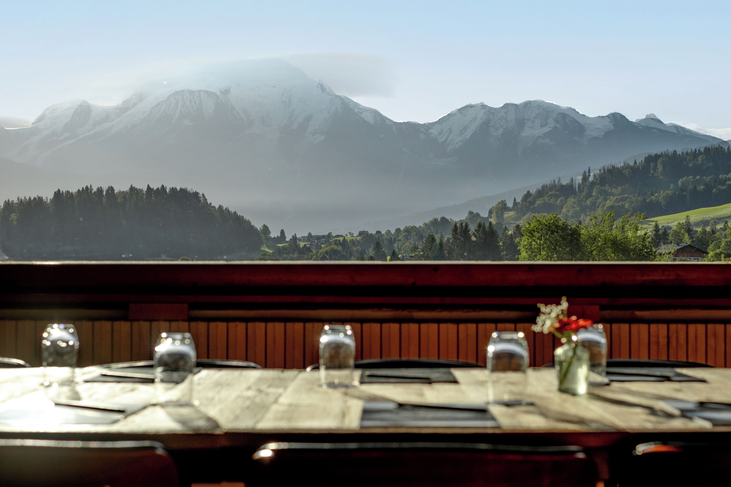 A restaurant table with a mountain view, wooden benches, and a vase with flowers.