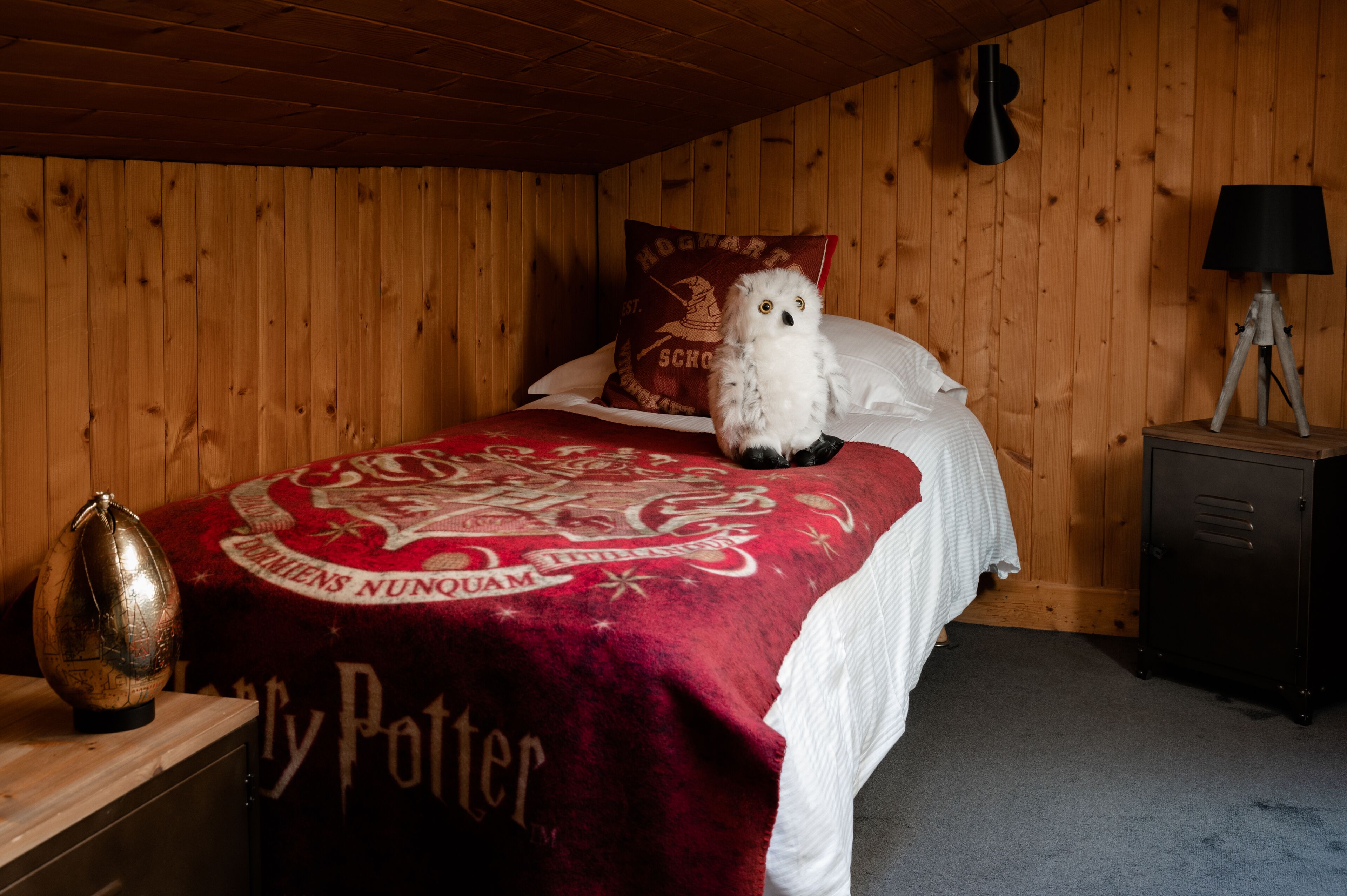 Tunnelmallinen huone, jossa on puuseinät, sänky, jossa on Harry Potter -teemainen peitto, lamppu puisen yöpöydän päällä ja koriste-esine lipaston päällä.