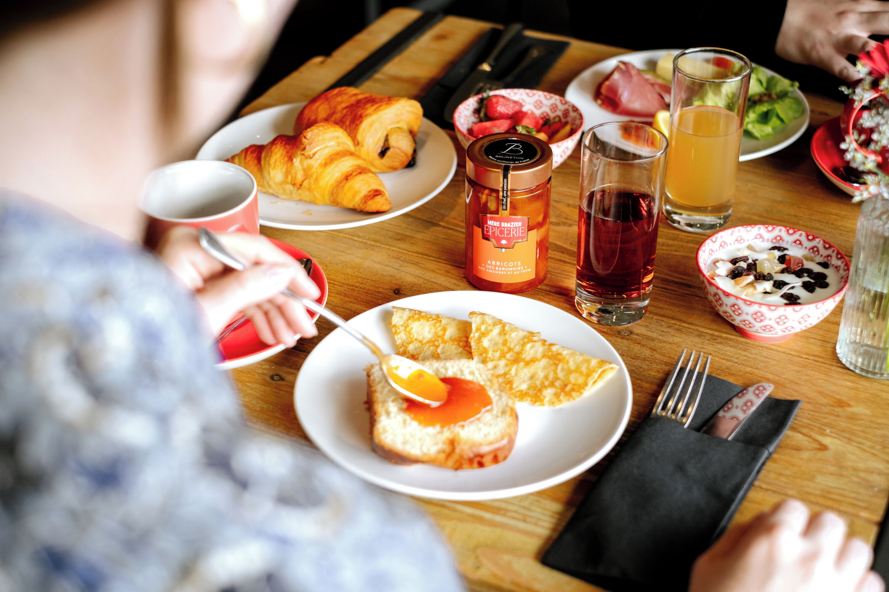 A breakfast table with croissants, a jar of jam, a glass of juice, and a bowl of fruit.