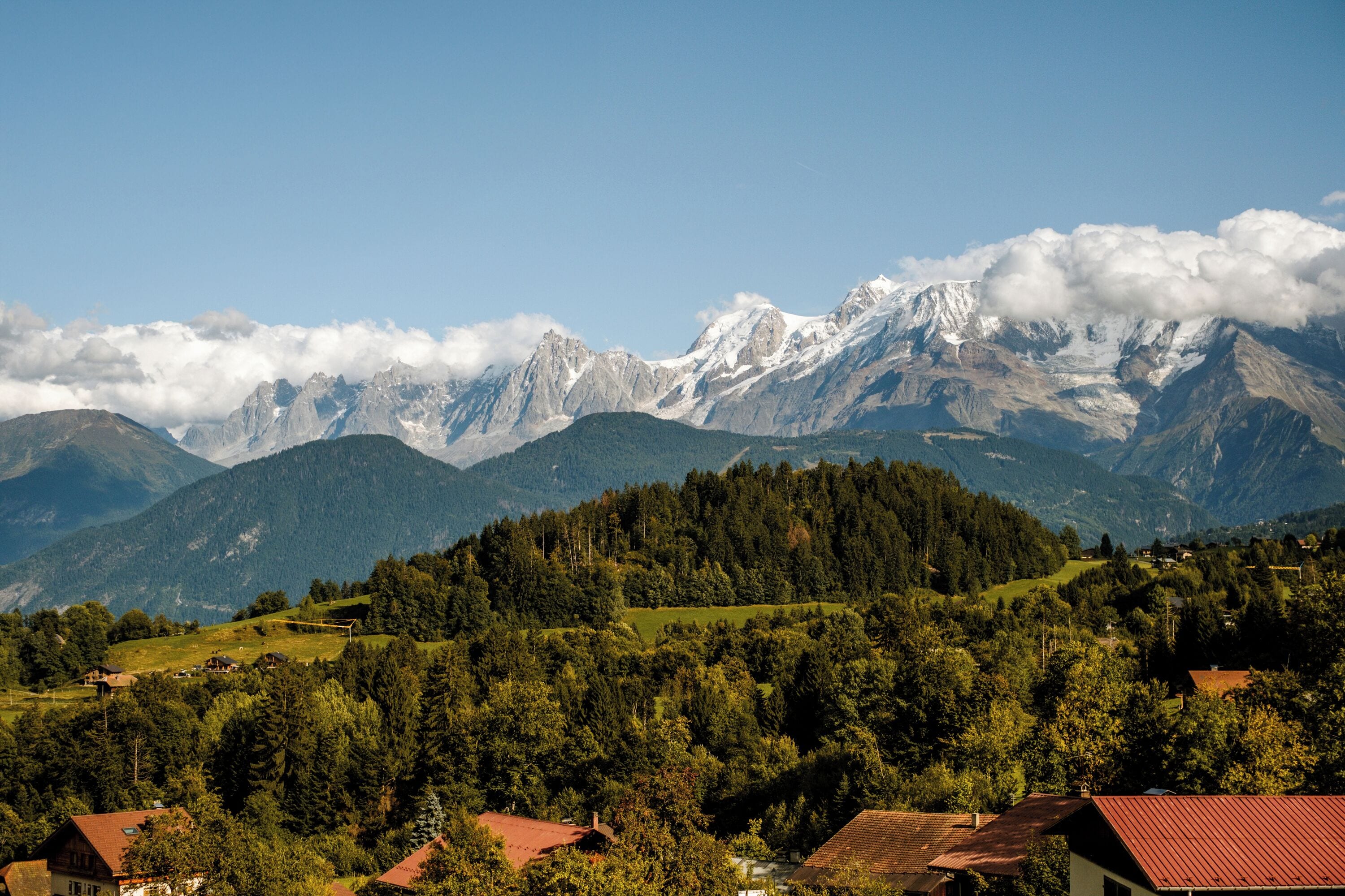 Un paysage montagneux avec des sommets enneigés, une vallée boisée et plusieurs bâtiments aux toits rouges.