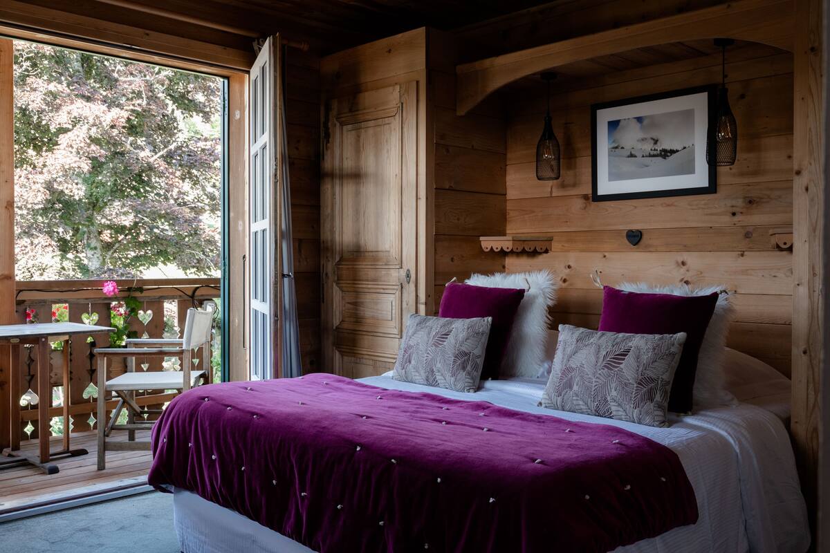 A wooden cabin bedroom with a bed, pillows, a framed picture, and a window overlooking greenery.