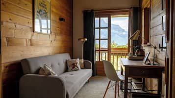 Ein gemütliches Zimmer mit Holzvertäfelung, einem grauen Sofa mit gemusterten Kissen, einem weißen Sessel, einem Schreibtisch mit Wasserkocher und Blick auf Berge durch ein Fenster.
