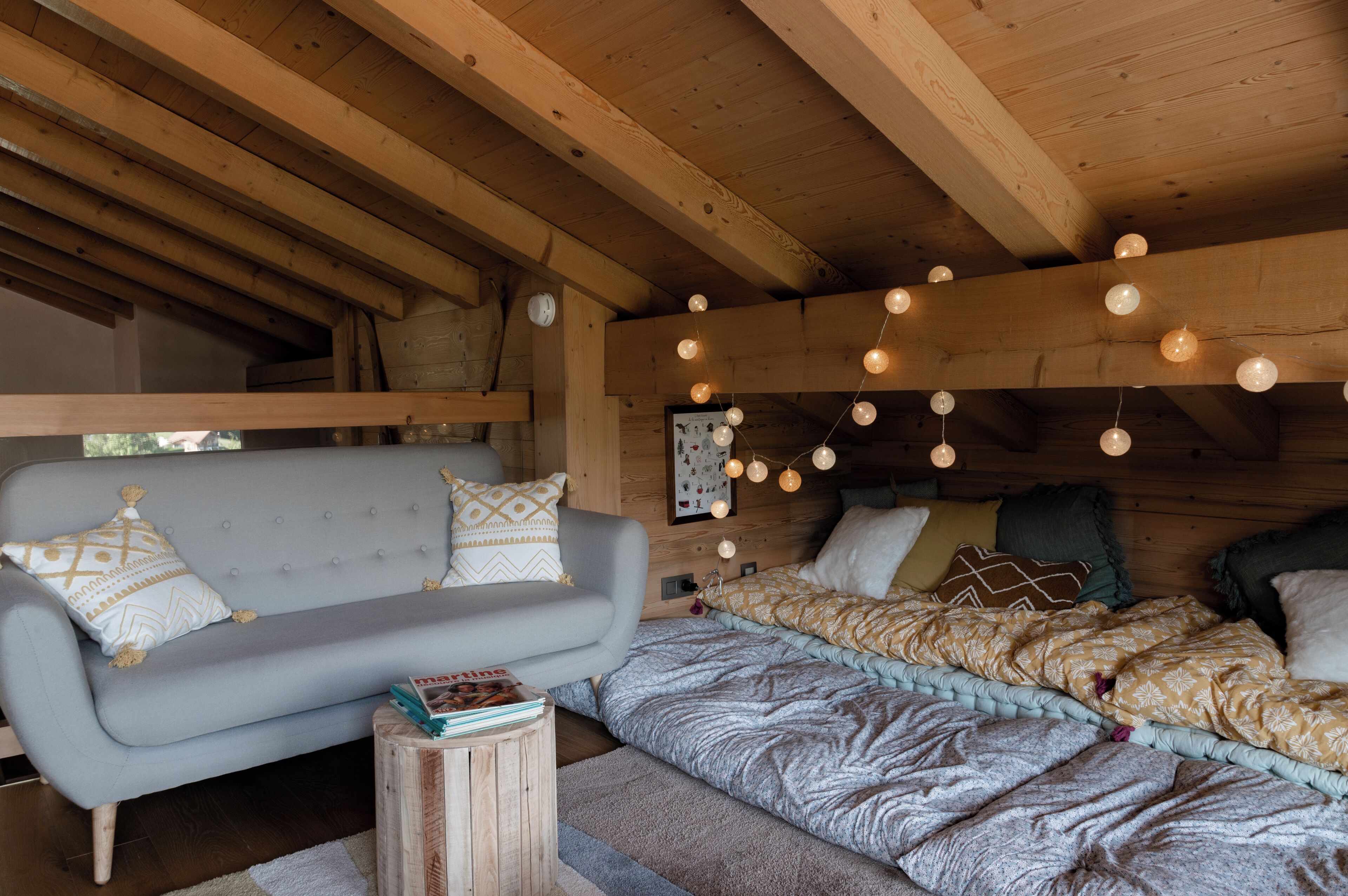 Interior acogedor de una cabaña con sofá, cama con ropa de cama estampada y techo de madera con luces colgantes.