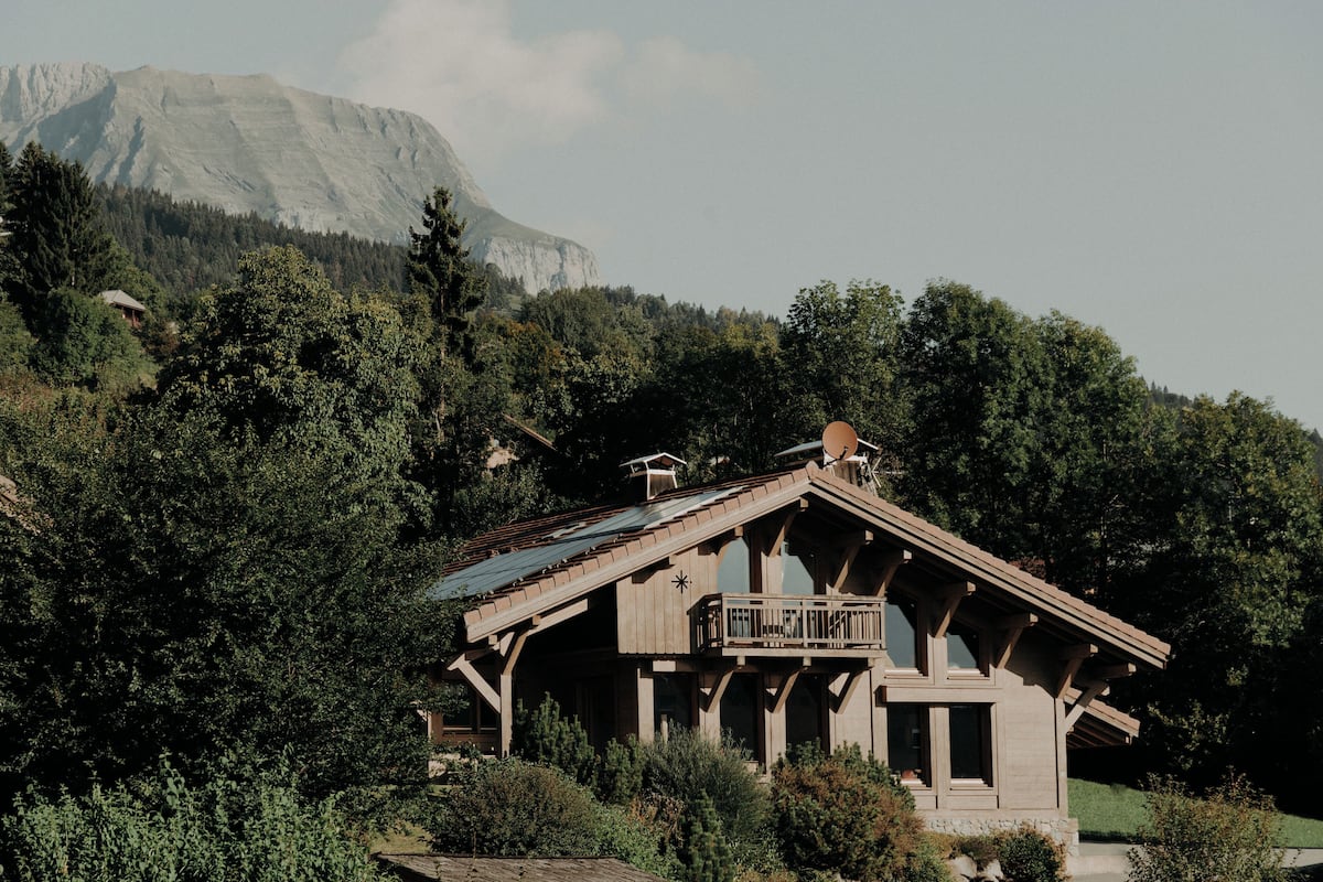 A wooden cabin with a balcony, surrounded by dense forest and a mountain in the background.