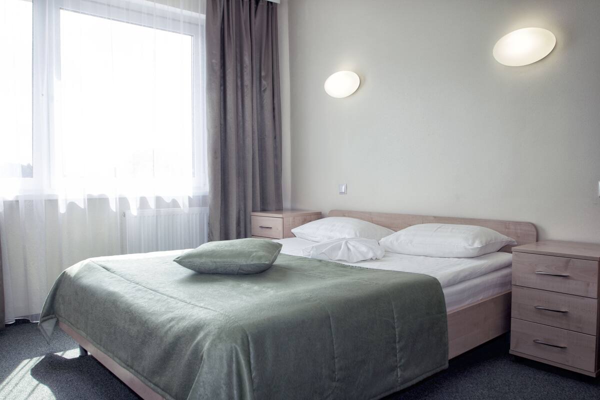 A neatly made bed with a green bedspread and pillows, a bedside table with drawers, and a wall-mounted light fixture.