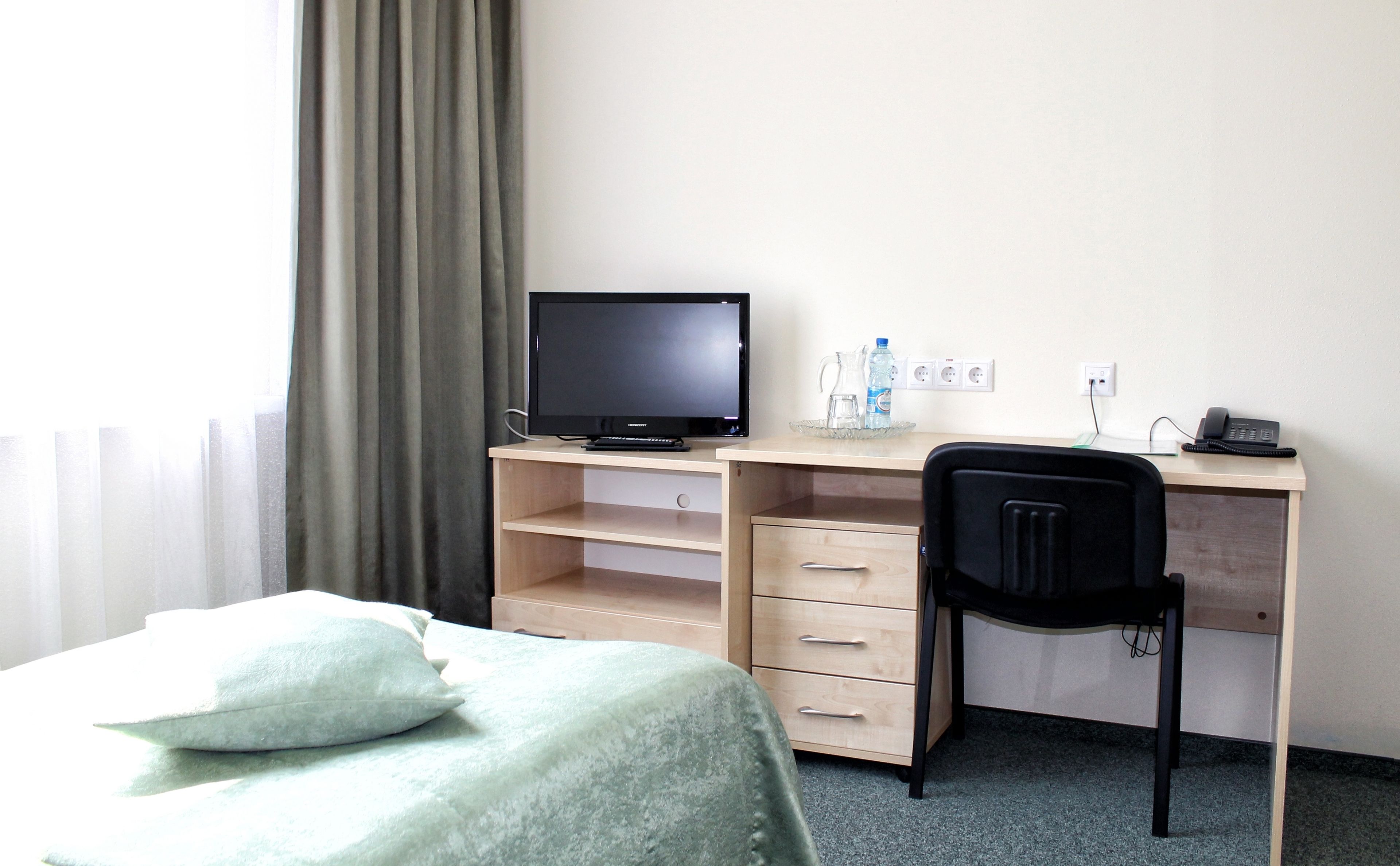 A hotel room with a bed, a desk with a computer, a chair, and a window with curtains.