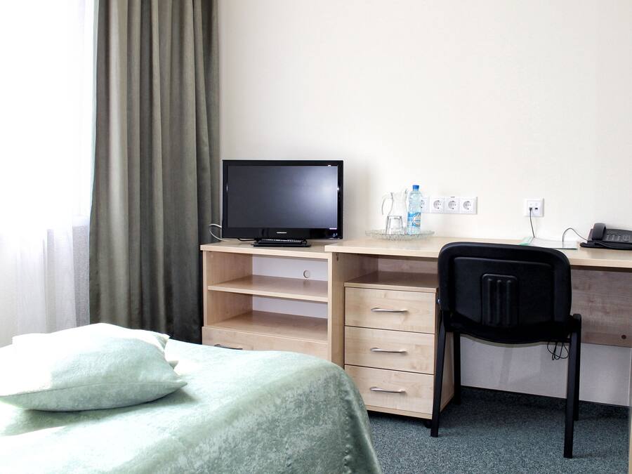 A hotel room with a bed, a desk with a computer, a chair, and a window with curtains.