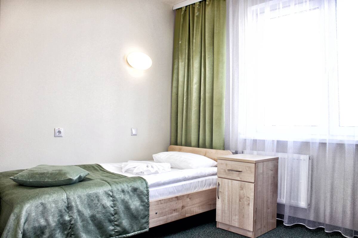 A single bed with a green blanket and pillow, a wooden nightstand with a drawer, a wall-mounted light, and a window with sheer curtains.