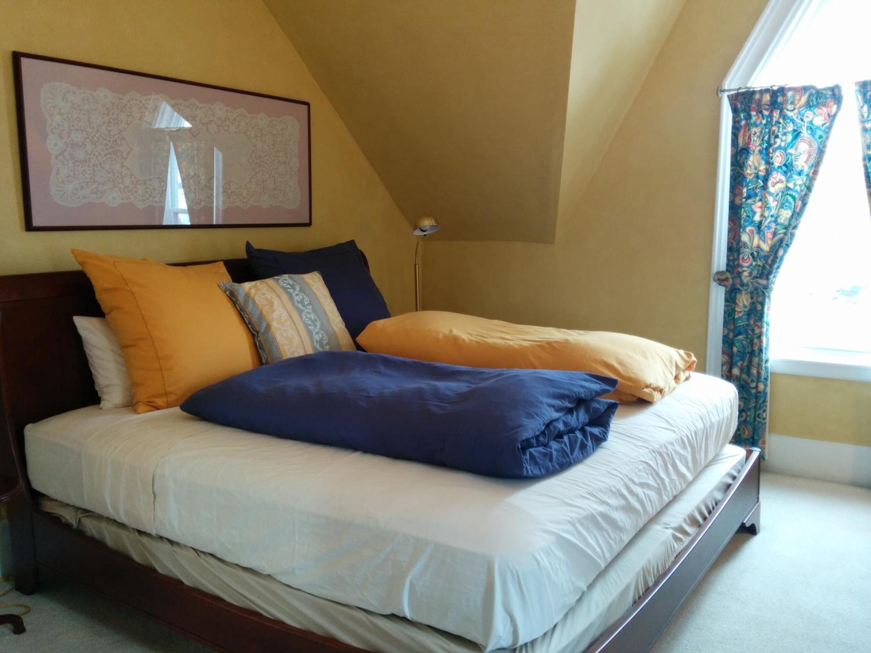 Een net opgemaakt bed met meerdere kussens in een kamer met een schuine kap en een groot raam met patroon gordijnen.