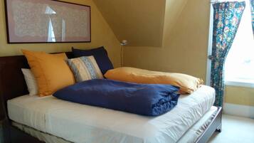 Een net opgemaakt bed met meerdere kussens in een kamer met een schuine kap en een groot raam met patroon gordijnen.