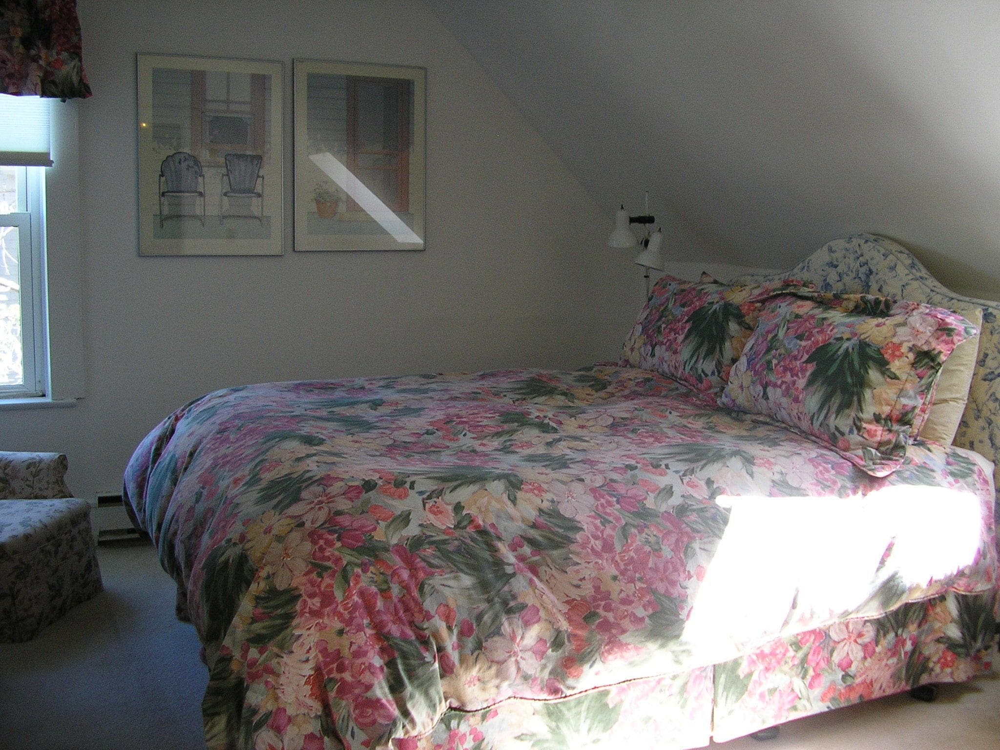 A bedroom with a floral bedspread, a nightstand with a lamp, and two framed pictures on the wall.