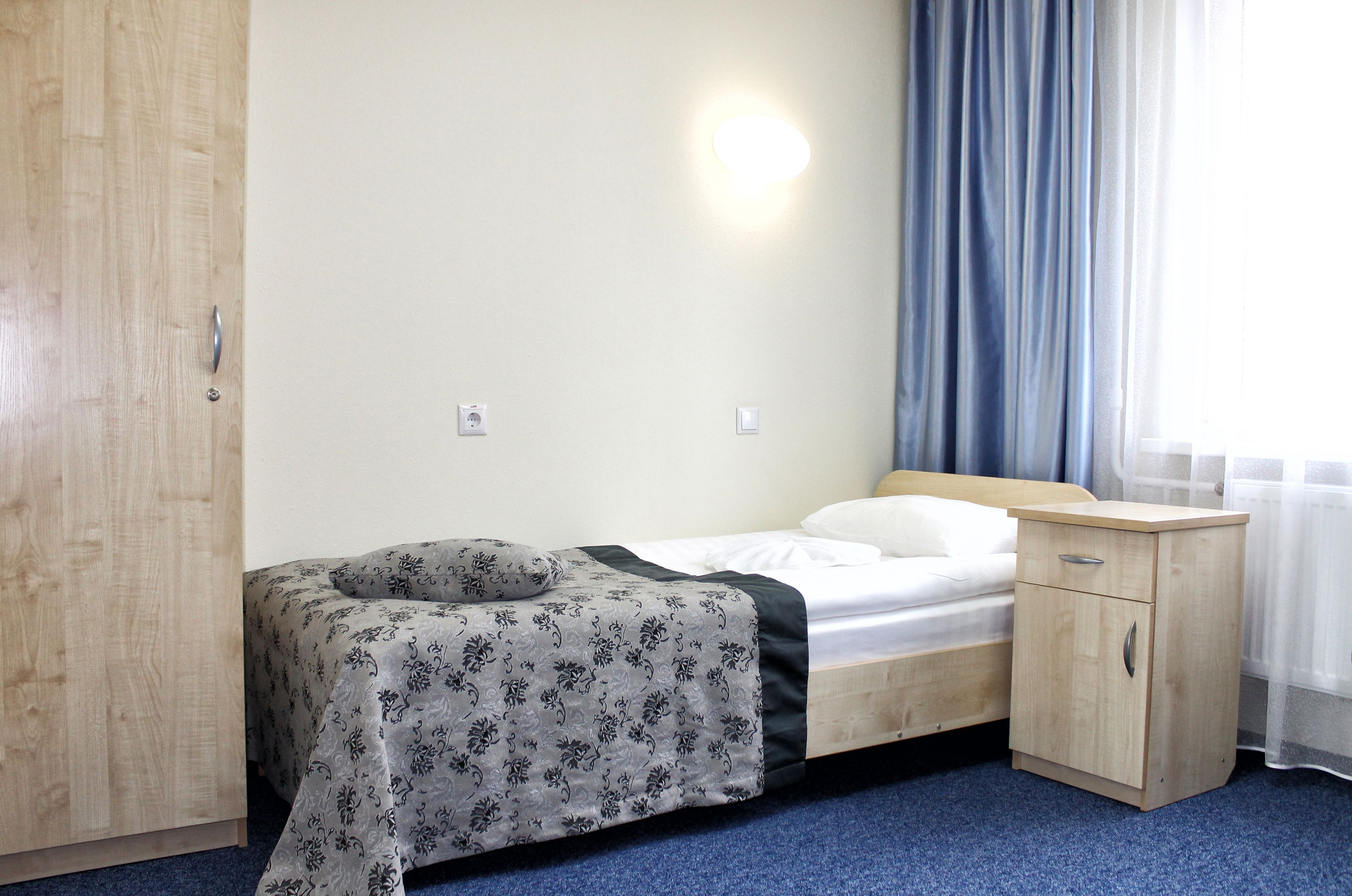 A single bed with a floral bedspread, a wooden nightstand, a wardrobe, and a window with curtains.
