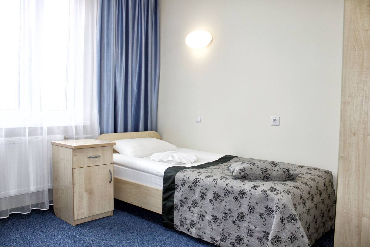 A small, neatly arranged bedroom with a single bed, a bedside table, and a wall-mounted light.