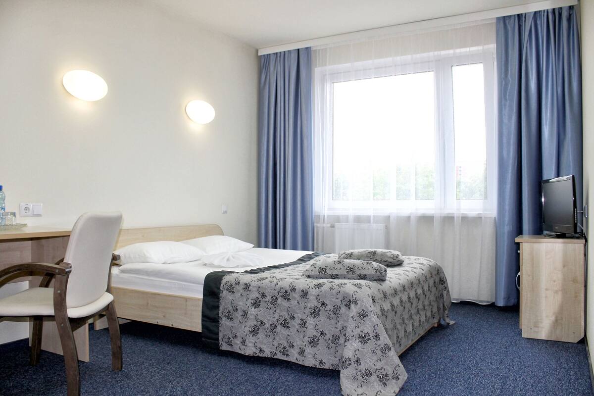 A hotel room with a bed, a desk with a chair, a television, and a window with curtains.