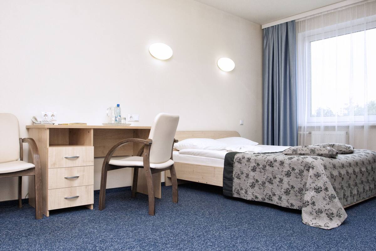 A hotel room with a wooden desk, a chair, a bed with a floral bedspread, and a large window with curtains.