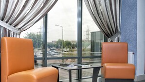 Un hall d’hôtel moderne avec deux fauteuils en cuir orange, une petite table ronde et de grandes fenêtres donnant sur le paysage urbain.