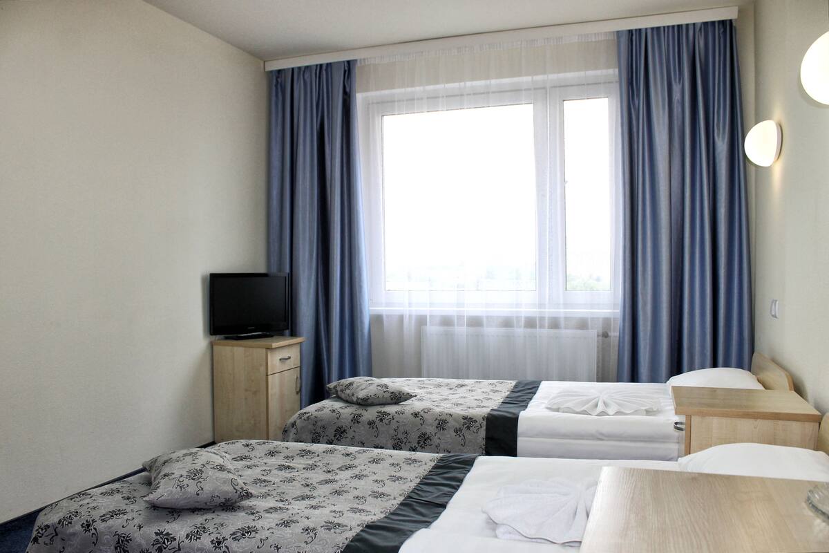 A hotel room with two beds, a TV on a wooden cabinet, and a window with blue curtains.